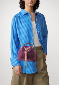 Piccola borsa viola con paillettes e chiusura a coulisse, tracolla a catena dorata, tenuta in mano, indossata con una camicia a righe blu e bianche.