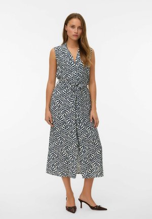 Mujer de pie con vestido sin mangas hasta la rodilla con patrón de hojas en azul y blanco y cintura anudada, combinado con tacones oscuros y puntiagudos.