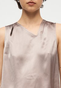 Κοντινό πλάνο ατόμου που φοράει αμάνικο, γυαλιστερό μπεζ σατέν τοπ με V-λαιμόκοψη σε απλό ανοιχτό φόντο.