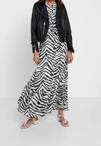 Svart läderbikerjacka över en vit och svart zebra-mönstrad maxiklänning med en lager-på-lager kjol, matchad med vita ankelrem-sandaletter med klack.