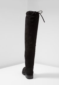 Botas negras de ante por encima de la rodilla, con una forma ajustada, puntera redonda y un lazo decorativo en la parte superior. Suela texturizada baja.