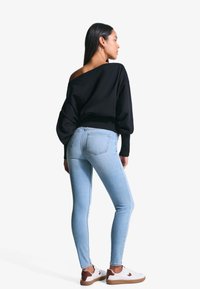 Crni oversized pulover, svijetloplave uske traperice, bijele tenisice s smeškastim akcentima, glatka tekstura, opušteni kroj. Model prikazan sa leđa.
