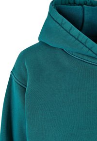 Tirkīza krāsas kokvilnas hoody ar mīkstu tekstūru, ar lielu divkāršu kapuci un redzamām šuvju detaļām ap kaklu.