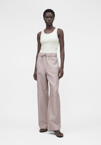 Μαύρη γυναίκα που φοράει λευκό αμάνικο τοπ, ροζ φαρδιά παντελόνια με κορδόνι και τσέπες, και μαύρα σανδάλια με ανοιχτά δάχτυλα, στέκεται σε λευκό φόντο.