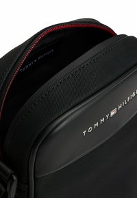Bolso negro de Tommy Hilfiger con un exterior texturizado, cremallera roja y detalles de logotipo. La abertura tiene un interior suave y forrado.