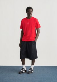 Άνδρας που στέκεται σε μπλε χαλί, φορώντας κόκκινη μπλούζα, μαύρα μακριά σορτς, άσπρες κάλτσες και μαύρα αθλητικά παπούτσια Nike, κοιτώντας προς τα αριστερά του.