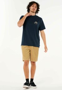 Marineblå bomulds T-shirt med frontlogo design, parret med lysebrune corduroy-shorts. Modellen bærer sorte sneakers og ankelstrømper.
