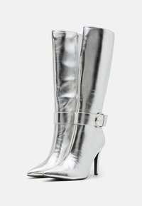 Stříbrné kozačky až po kolena se špičatou špičkou, hladkou kovovou texturou a širokým páskem s kulatou přezkou. Vysoký podpatek.