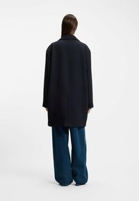 Svart yllekappa med notch-krage och oversize passform. Kombineras med lösa blå jeans, vilket visar på en minimalistisk design och en jämn textur.