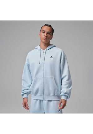 Junger Mann mit geflochtenem Haar, der einen hellblauen Kapuzenpullover mit Reißverschluss und passende Jogginghose mit schwarzem Jumpman-Logo auf Brust und Bein trägt.