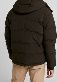 Mørkebrun pufferjakke med hætte, der har en diagonal quiltet mønster og elastiske manchetter, båret over lyse bukser.