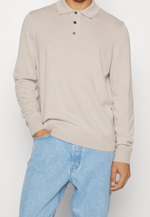Beige Langarm-Polohemd mit weicher Textur, Knopfleiste mit drei Knöpfen und gerippten Bündchen, kombiniert mit hellblauen Jeans.
