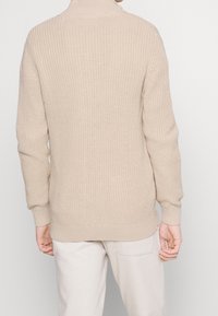 Beżowy sweter z dzianiny z półwysokim kołnierzem, teksturowanym wzorem oraz prążkowanymi mankietami i dołem. Noszony na jasnych spodniach, prezentujący luźny krój.