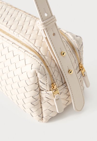 Τσάντα από κρεμ πλεγμένο δέρμα με χρυσά φερμουάρ και ρυθμιζόμενο λουράκι με χρυσά καρφιά, εμφανιζόμενη σε κοντινή λήψη.