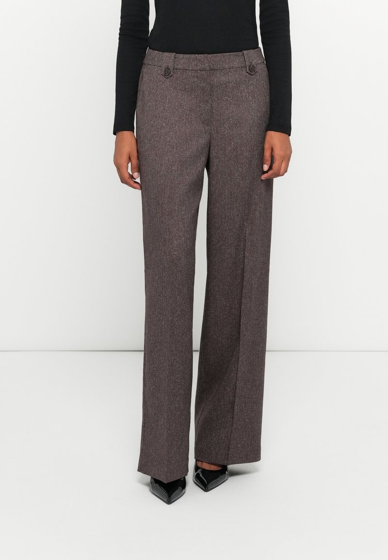 Personne portant un pantalon gris taille haute à jambes larges avec des détails de boutons, un haut noir à manches longues et des chaussures noires pointues, debout sur un sol blanc.