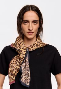 Sciarpa leopardata gialla e marrone con bordi sfilacciati, indossata attorno al collo; abbinata a una maglietta nera. Tessuto texturizzato.