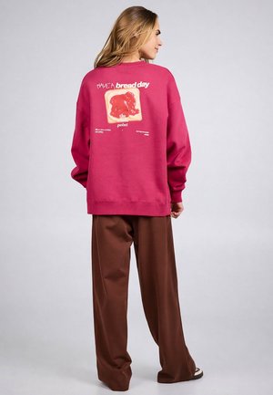 Vrouw draagt een roze sweatshirt met een toast en de tekst "Have a bread day" op de achterkant, gecombineerd met losse bruine broek en casual schoenen.