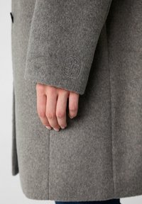 Hand ligt naast een grijs wollen jasje met een klein rond logo geborduurd nabij de manchetten.