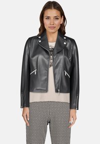 Czarna kurtka motocyklowa z ekologicznej skóry z srebrnymi elementami, zapinana na zamek, z kieszeniami na zamek i klasycznym kołnierzem, noszona na beżowej bluzce i wzorzystych spodniach.