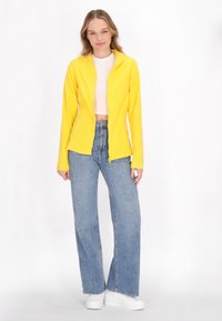 Giacca a zip gialla con cappuccio, maniche lunghe, abbinata a jeans a gamba larga blu chiaro e sneakers bianche. Tessuto liscio con un design aderente.