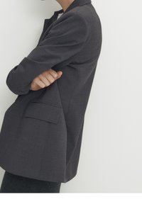 Blazer grigio scuro con una finitura texturizzata, taschino sul petto singolo, revers a punta e una vestibilità leggermente ampia, mostrato su uno sfondo neutro.