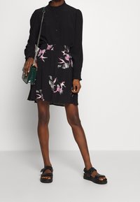 Blusa nera con colletto a volant e chiusura con bottoni abbinata a una gonna nera con motivi floreali rosa e pois argento. Sandali neri e borsa verde.