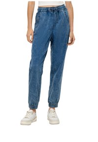 Modré džínové jogger kalhoty s elastickým pasem, nastavovací šňůrkou, manžetami na kotnících a volným střihem. V kombinaci s bílými teniskami.