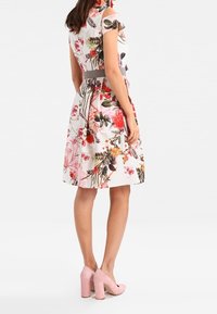 Donna che indossa un vestito bianco alla lunghezza del ginocchio con motivi floreali rosa e verdi, abbinato a scarpe con tacco a blocco rosa, in posizione laterale.