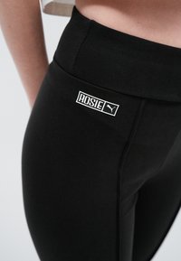 Krupan prikaz crnih sportskih tajica s visokim strukom, s bijelim natpisom "ROSIE" i Puma logotipom na bokovima.