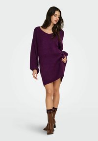 Fioletowa, dziergana, oversize sukienka-sweter z szerokim dekoltem i długimi rękawami, z bocznym rozcięciem. Noszona z brązowymi kozakami za kolano.