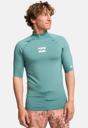 Krótki rękaw, turkusowa koszulka ochronna z wysokim kołnierzem i białym logo fali na klatce piersiowej, wykonana z gładkiego, elastycznego materiału.