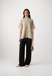 Beige blouse with short sleeves, featuring embroidered details and buttons down the front. Paired with black wide-leg pants and a small bag.