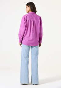 Rosa Hemd mit Knopfleiste und langen Ärmeln, lockere Passform, mit seitlichen Falten im Rückenteil, kombiniert mit hellblauen Schlagjeans, vor einem neutralen Hintergrund.