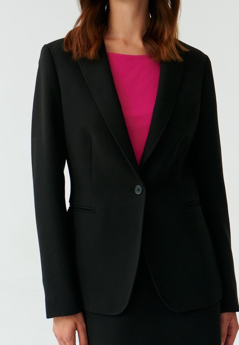 Femme portant un blazer noir cintré sur un haut rose vif, vue du cou aux hanches sur un fond clair uni.