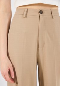 Béžové kalhoty s vysokým pasem vyrobené z hladké tkaniny, se zapínáním na jeden knoflík, bočními kapsami a přizpůsobeným střihem.