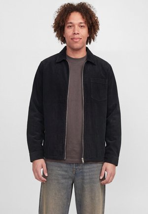Un homme aux cheveux bouclés se tient devant un fond clair. Il porte une veste noire zippée, un t-shirt gris et un jean délavé.
