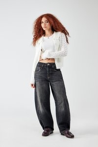Vrouw met lang krullend rood haar, gekleed in een witte gebreide cardigan, witte crop top, wijde zwarte jeans en donkere loafers tegen een eenvoudige achtergrond.