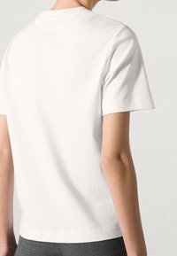 White cotton T-shirt with short sleeves, a rounded neckline, and a subtle texture, viewed from the back. No visible graphics or logos.