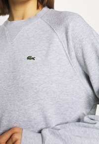 Sudadera gris hecha de tela suave, con cuello en V, mangas raglán y un pequeño logo de cocodrilo verde en el pecho.
