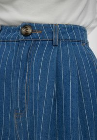 Kék farmer magas derekú nadrág fehér függőleges csíkokkal, elülső gombos záródással és varrt ráncokkal a derékvonal mentén.