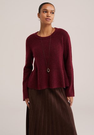Femme portant un pull bordeaux à manches longues avec une jupe plissée marron et un long collier avec un pendentif circulaire.
