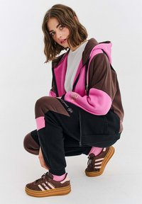 Młoda kobieta z brązowymi włosami, ubrana w brązowy, różowy i czarny dres oraz brązowe buty Adidas, klęczy na jednolitym białym tle.