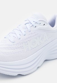 Scarpa sportiva bianca con tomaia textured, suola spessa e ammortizzata, e logo HOKA ben visibile. Presenta lacci piatti e fori di ventilazione laterali.
