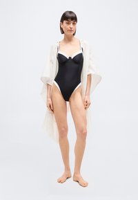 Γυναίκα που στέκεται ξυπόλητη φορώντας μια ασπρόμαυρη ολόσωμη μαγιό με λευκή κάλυψη με λεπτομέρειες κρόσι.