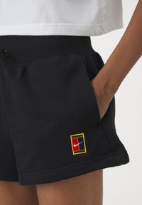 Calções de desporto pretos feitos de tecido suave, com um emblema da Nike com borda amarela e formas geométricas em vermelho e azul no bolso esquerdo.