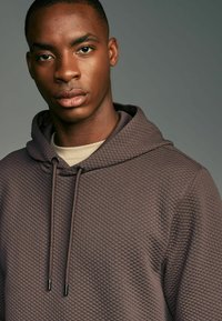Brun quilted hoodie med snor i hætten, der har et struktureret mønster og en rund halsudskæring. Laget over en beige skjorte.