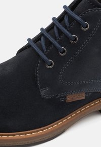 Gros plan d'une bottine en daim bleu marine avec des lacets bleus, des œillets métalliques, des coutures détaillées, et une semelle marron avec couture blanche.