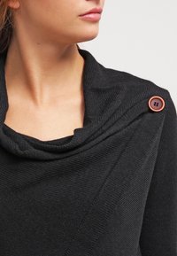 Pull noir en maille avec col cheminée, présentant une texture en côtes diagonales et un détail de bouton orange sur l'épaule.