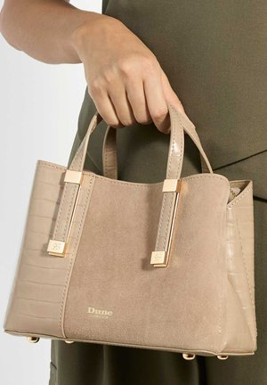 Mano sosteniendo un bolso de ante beige y piel sintética de cocodrilo con detalles dorados y el logo de Dune London, sobre ropa de color verde oliva.