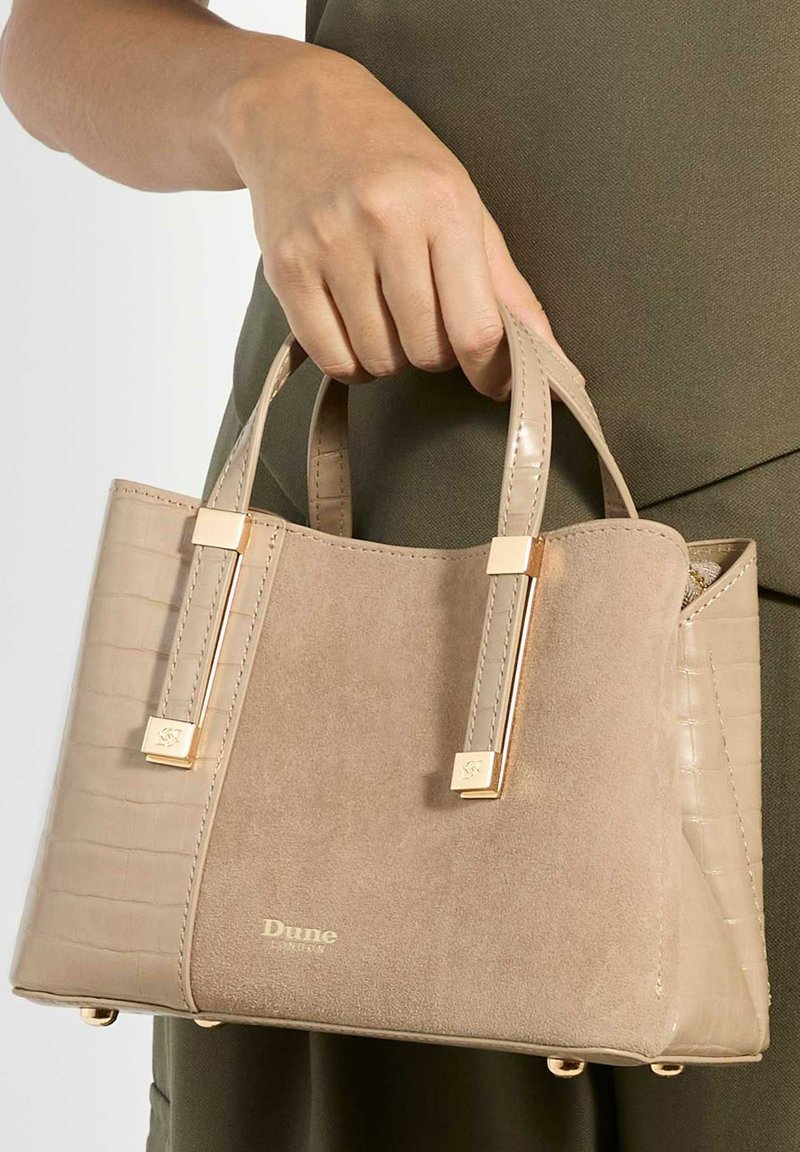 Main tenant un sac à main en daim beige et simili cuir croco avec des accents dorés et le logo Dune London, sur un vêtement vert olive.